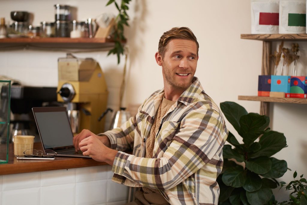 image | Manage Your Property Man in a plaid shirt working on a laptop at a cafe counter, smiling and looking to the side.