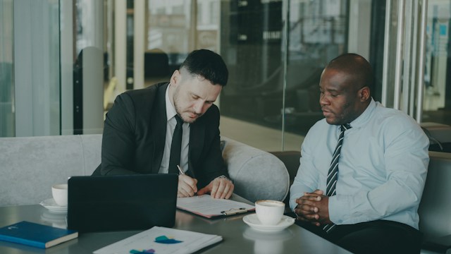 Sign a Short-Term Licence or Bridge Agreement | Manage Your Property Two businessmen in suits sit at a table with a laptop, reviewing a document as one writes notes and the other listens, coffee cups nearby.