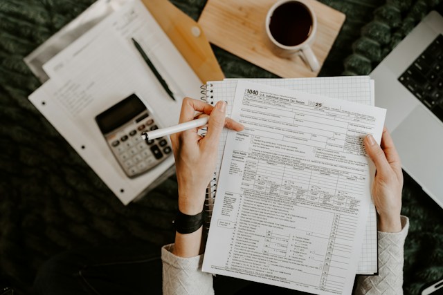 Who Really Pays Recovering Management Costs from Tenants | Manage Your Property Close-up of hands writing on a 1040 tax form with a pen, calculator, coffee cup, and laptop nearby on a desk.